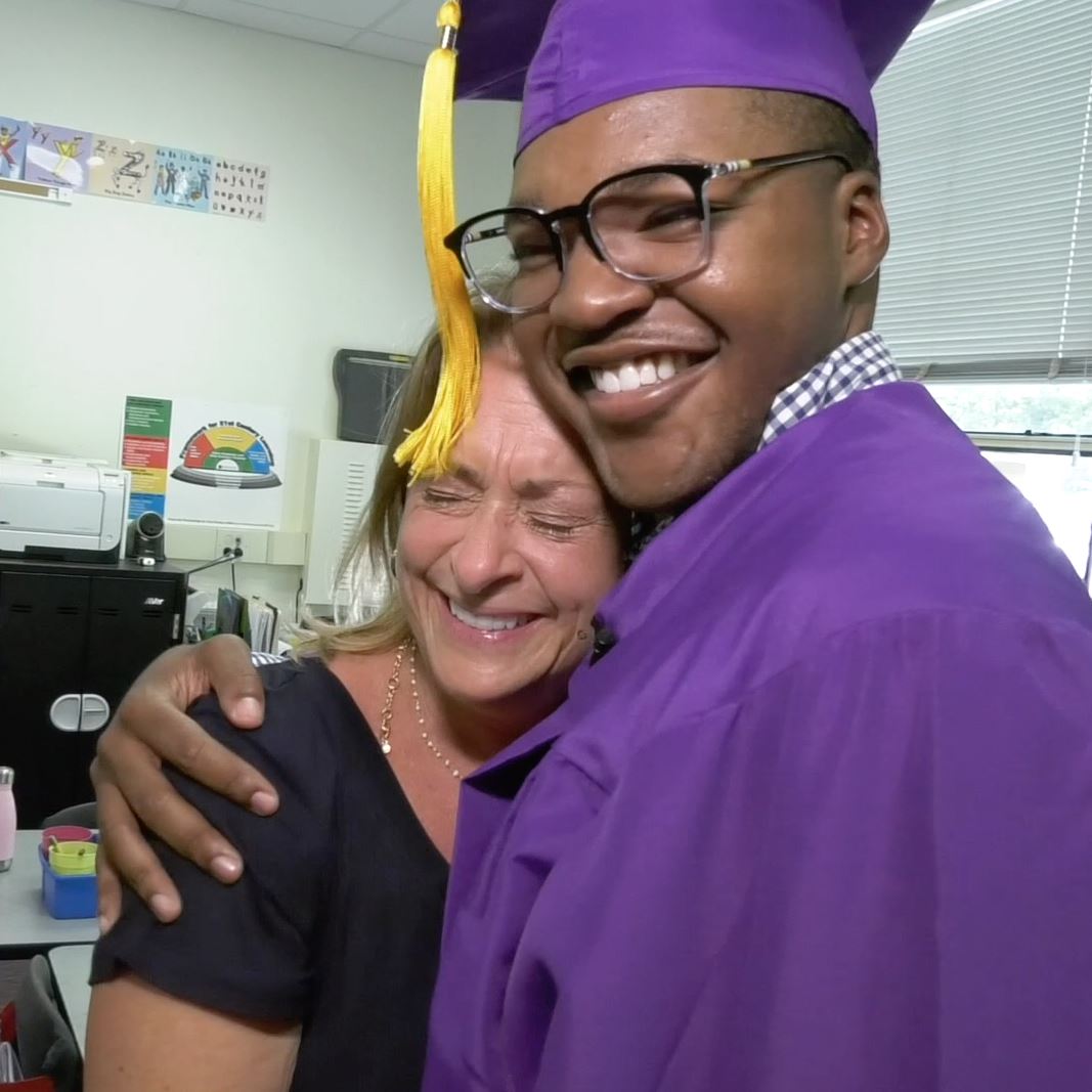  Senior wearing cap and gown, hugging his former kindergarten teacher